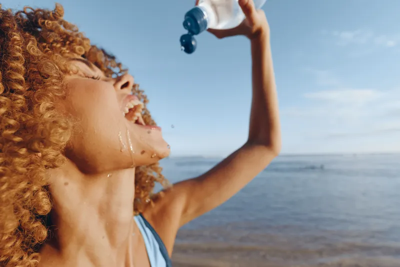 Young woman with curly hair drinks from a salt shaker on a sunny beach, the sea and sky shimmer behind her, a spontaneous outdoor moment