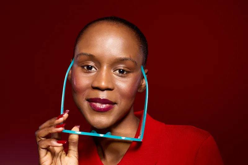 Close-up portrait of a smiling African woman with dark skin holding blue eyeglasses against a dark red background. Concept of beauty, confidence, and fashion accessories for women.