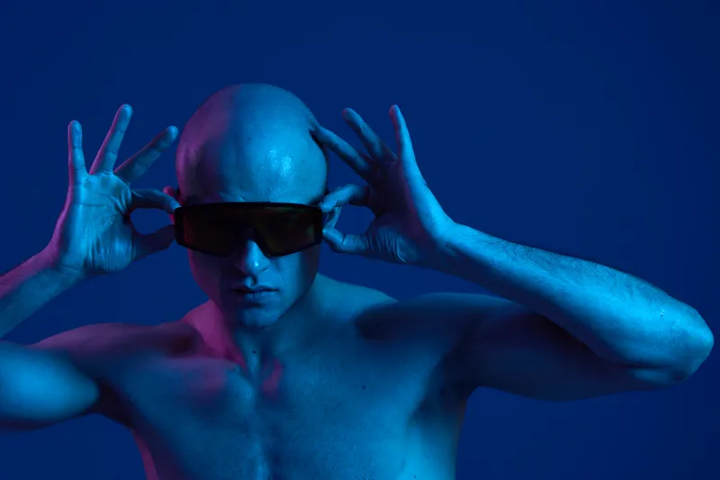 A man with a bald head wearing dark sunglasses poses dramatically against a vibrant blue background, showcasing an expressive and artistic composition in this color image.