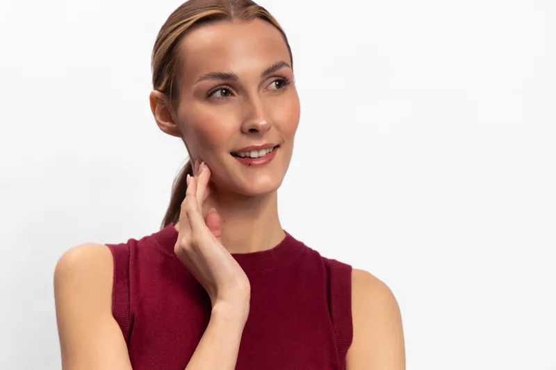 Young woman with clear skin and light makeup looks away smiling gently, wearing sleeveless burgundy top. Portrait on white background with soft lighting creates calm and positive mood.