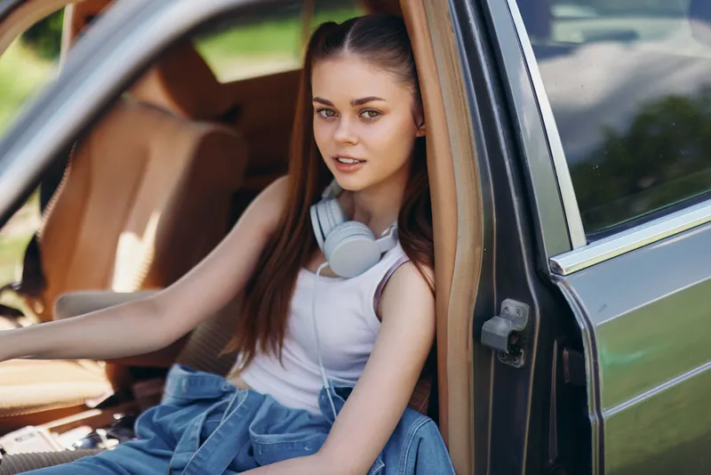 Young woman relaxing in a car wearing headphones, denim jacket, and a casual tank top, surrounded by a bright green natural background, evoking a carefree summer vibe.