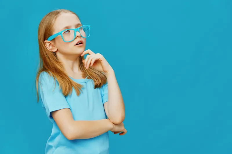 Thoughtful girl with blue glasses on a turquoise background, wearing a light blue shirt, expressing curiosity and contemplation with a playful vibe