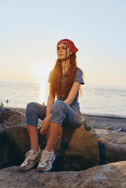 Lifestyle woman in modern boho-western denim street style sitting on rocks by the sea at sunset with a red bandana and casual sneakers, film color effect.