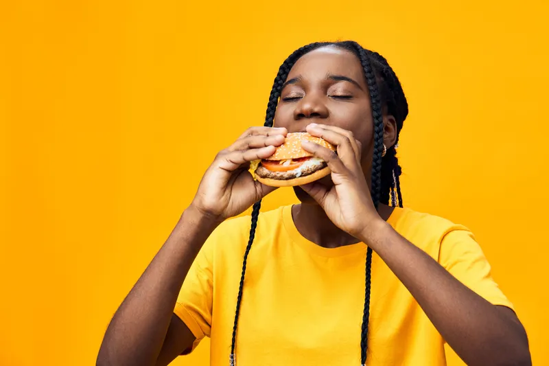Young African woman enjoying a flavorful burger against a vibrant yellow background, showcasing happiness and enthusiasm for delicious fast food