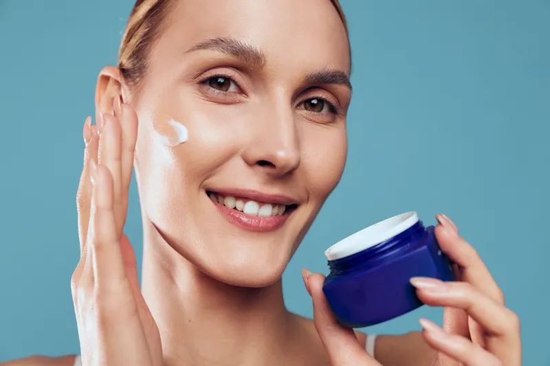 Cheerful woman applying moisturizer to her face, close up, healthy skin, soft lighting, skincare routine moment, calm atmosphere in background today