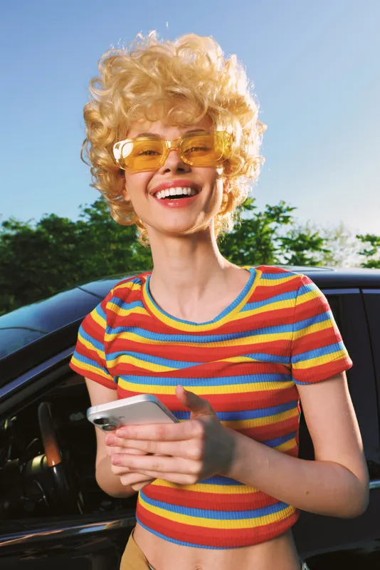 Smiling woman with curly blonde hair wearing a colorful striped crop top and yellow sunglasses, holding a smartphone beside a car outdoors. Retro film color mood, vibrant summer vibe.