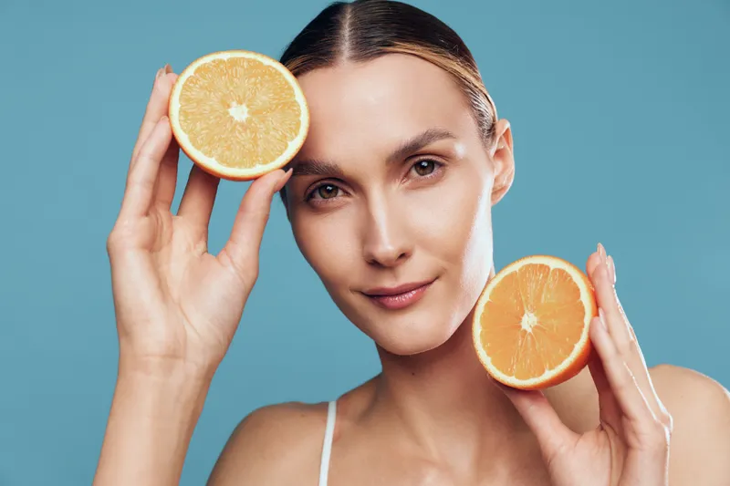 Woman beauty and skincare concept, holding orange slices near her face, vitamin C focus, calm expression, clean minimal studio background, wellness nutrition and healthy lifestyle