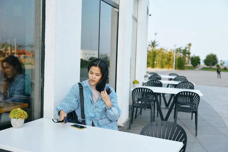 Woman sits at a cafe table beside a window outdoors, adjusting sunglasses while a phone rests on the table, empty chairs around, serene daytime scene for casual urban break, soft light highlights her