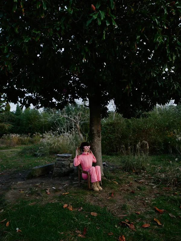 Young girl sits on a bench beneath a large tree in an outdoor park, wearing a pink coat. A calm autumn scene highlighting solitude, nature, and quiet reflection in a peaceful portrait.