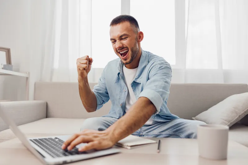 Excited man celebrating success while working on a laptop in a bright, modern living room. Relaxed atmosphere enhanced by natural light and stylish decor.