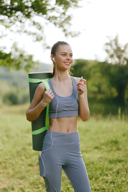 Young woman in activewear with a yoga mat walking in a green park, smiling and enjoying the fresh air, showcasing a healthy lifestyle in a natural setting