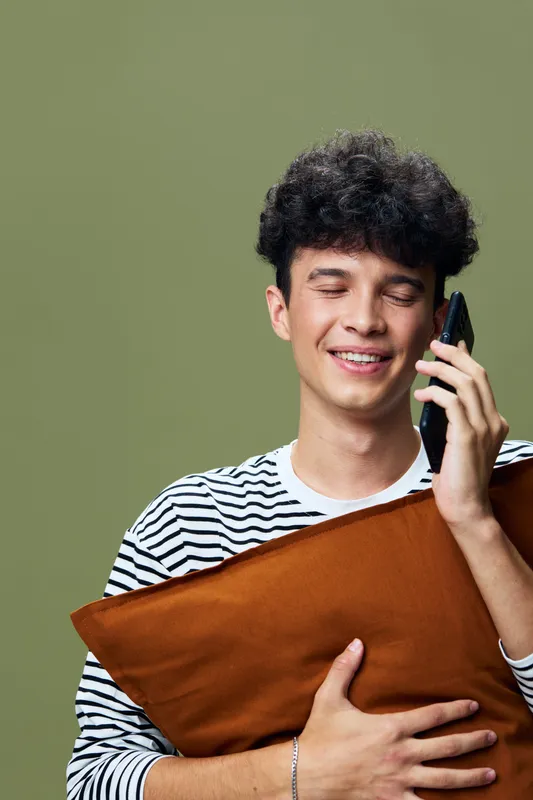young man smiling while talking on phone, holding brown pillow with closed eyes against olive green background, casual striped shirt expressing happiness and relaxation