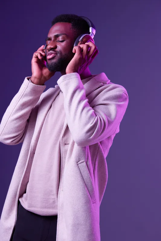 Young African man enjoying music with headphones against a vibrant purple background, dressed in a stylish beige coat, showcasing relaxed emotions and modern style