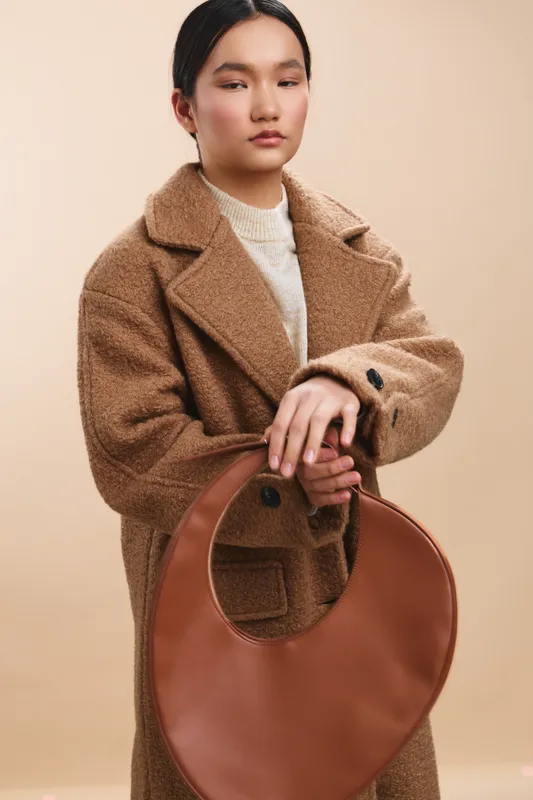 asian woman in a stylish coat stands with a large brown bag in a minimalist studio. her calm expression and poised posture convey elegance and confidence, creating a chic fashion moment.