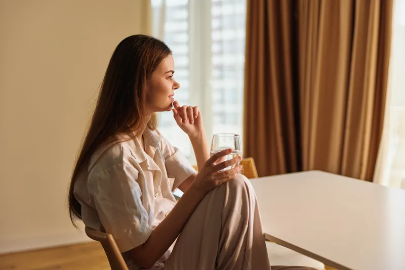 Woman sitting with a glass of water by the window at home, contemplative morning portrait with soft natural light, neutral interior, curtains, wooden chair, casual outfit and profile view.