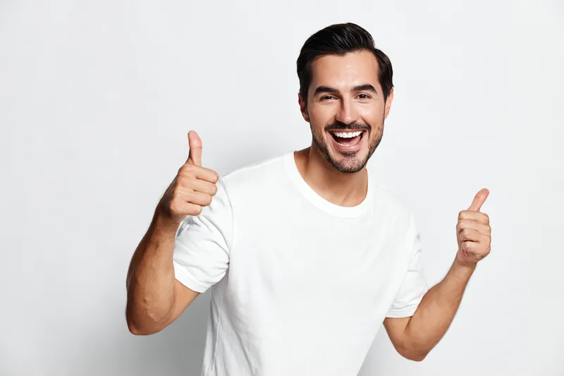 Happy smiling young caucasian man with short hair wears a white t-shirt, showing thumbs up with both hands against a plain light background. Positive lifestyle concept.