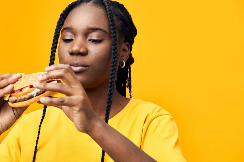 Young Black woman enjoying a delicious burger against a vibrant yellow background, showcasing joy and healthy eating habits