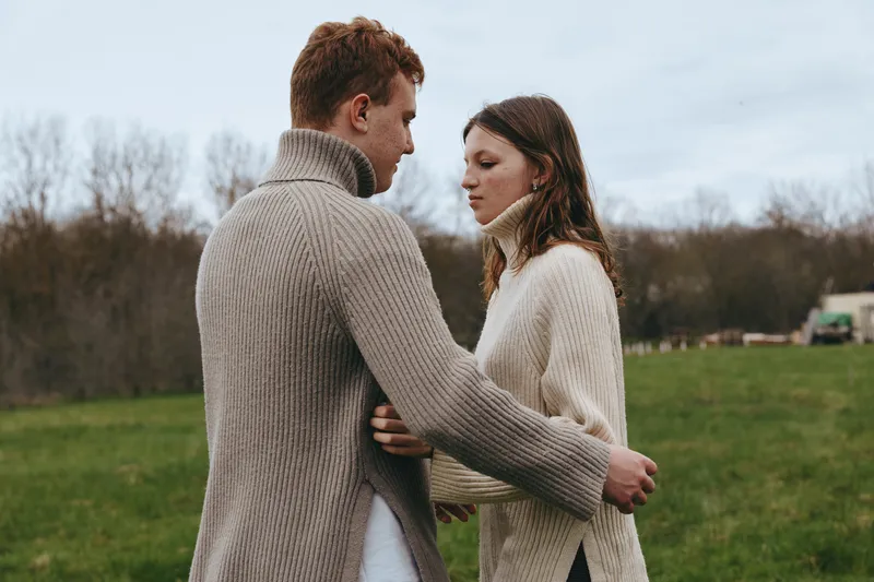 Couple in cozy sweaters share a quiet outdoor moment in a grassy field, embracing gently, under soft light, portraying warmth, connection, and a relaxed lifestyle scene.