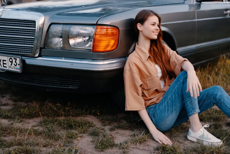 Young woman sitting next to a vintage car, wearing casual clothing, evoking a relaxed mood in a warm, natural setting during the golden hour.