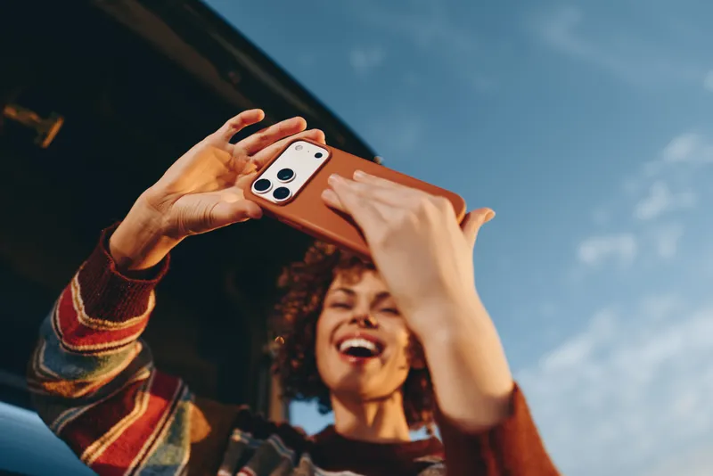 Woman lifestyle outdoors holding smartphone with hands, smiling and laughing, making a casual selfie, close foreground hands and phone, warm daylight, modern technology, blue sky background