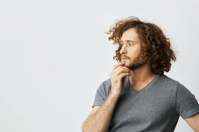 Pensive young man with curly hair contemplating against a neutral background, showcasing introspection and curiosity in casual gray attire