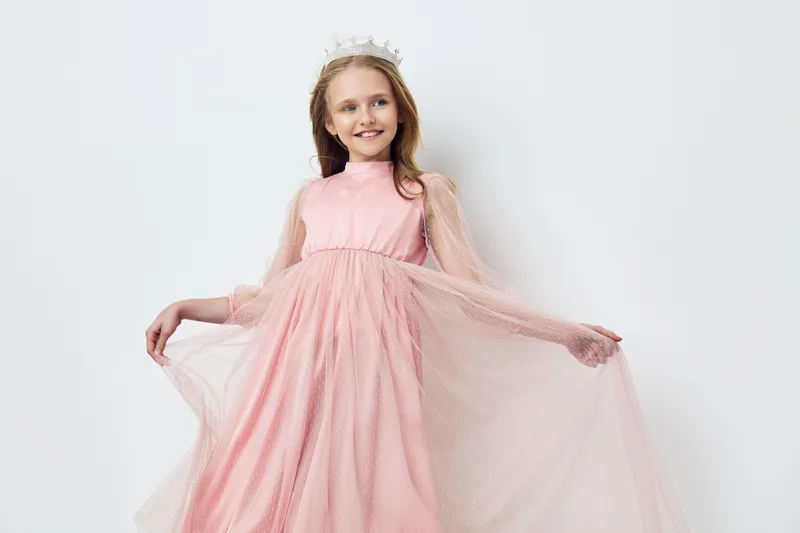 Smiling girl in a pink lace dress with a crown, showcasing joy and elegance in a soft pastel setting, perfect for celebrating special moments