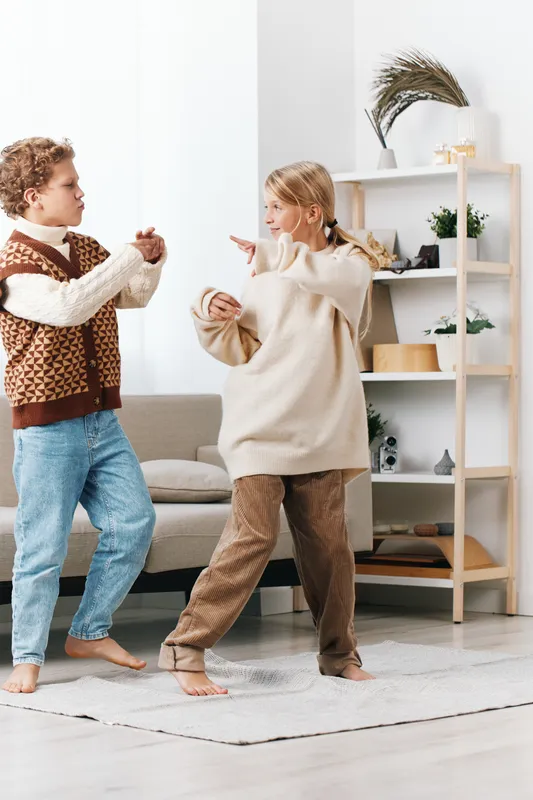 Two children dancing indoors in a bright living room, wearing cozy knitwear and warm tones, capturing a candid moment of playful movement on a soft rug.