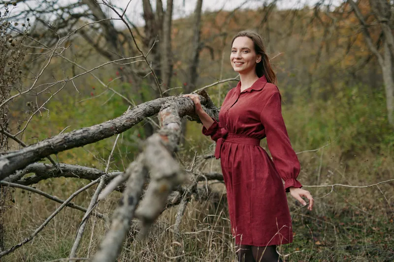 A smiling woman in a simple red dress stands beside a fallen tree in a grassy field, an authentic, credible candid portrait capturing natural light and grounded everyday beauty.