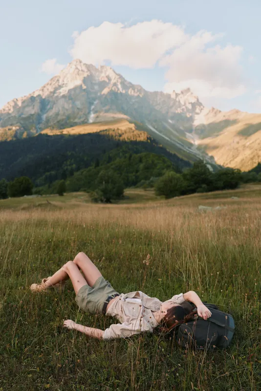 Young woman with brunette hair lies on a sunlit meadow near distant mountains, backpack nearby, enjoying a relaxed, carefree outdoor moment in the open field.