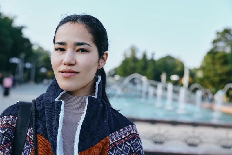 Woman in a patterned jacket standing outdoors near a park fountain, casual street style and confident mood, soft daylight, urban ambiance, fashion expression