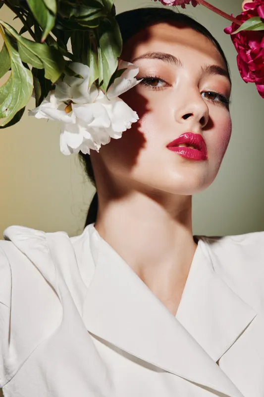 Close-up of a young woman with glossy red lipstick and smooth skin posing with flowers near her face. Artistic beauty fashion portrait with natural light shadows, creative makeup concept.
