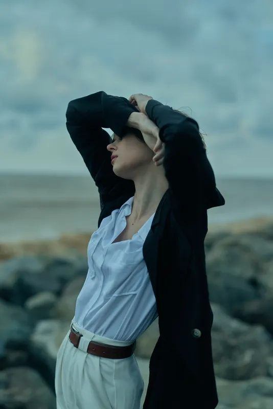 Woman on a windy beach in a blazer and white shirt, arms raised to frame the face as waves kiss the rocks, outdoor fashion mood, confident style blending corporate chic with coastal calm
