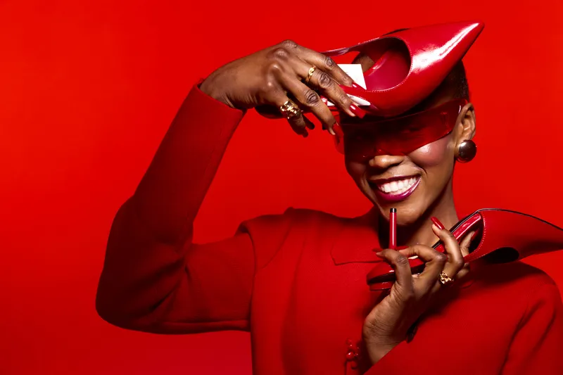 Smiling black woman with dark skin holds red high heel shoes near face on red background. Fashion concept with elegant accessories and joyful mood in studio portrait of African ethnicity.