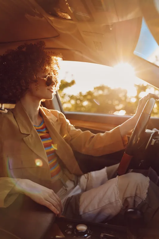 Cheerful woman driving a car at sunset with retro film color, striped top and blazer, sunglasses, and breeze in her hair, a candid lifestyle moment in warm natural tones.