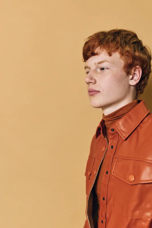 Young man with red hair and fair skin wearing a brown leather jacket and turtleneck with side profile on beige background. Stylish, casual, portrait and fashion concept
