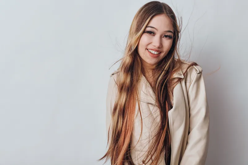Young woman with long hair wearing beige jacket and smiling against plain light background. Beauty, casual style, happy expression, portrait with natural light and positive mood.