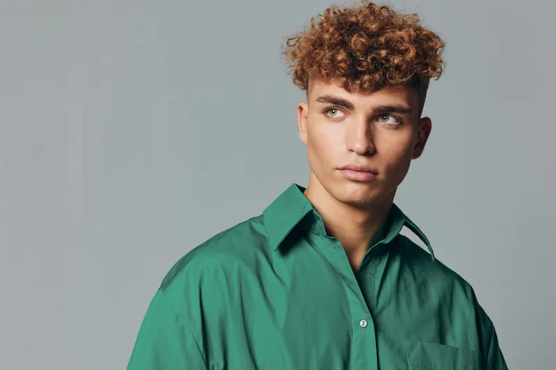 Young man with curly hair wearing green shirt looking sideways on gray background. Portrait of confident male model with casual style and thoughtful expression in studio lighting.