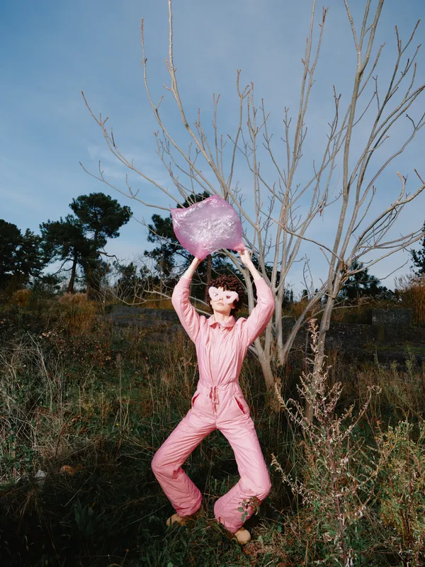 Person in a pink jumpsuit leaps outdoors, lifting a heart shaped object under a clear sky. Leafless trees and wild vegetation frame a playful, energetic scene perfect for lifestyle, wellness or