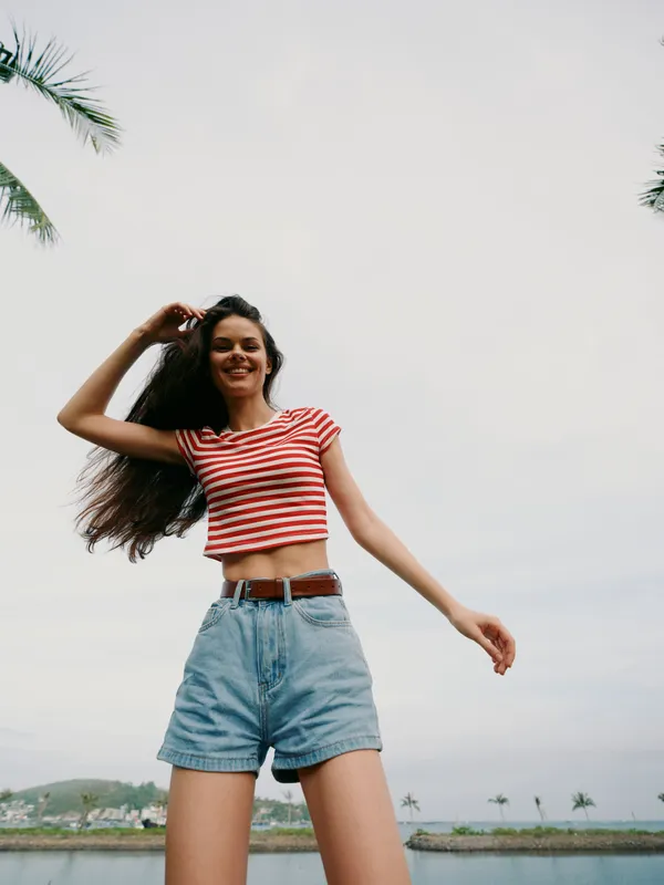 A joyful young woman poses outdoors in a striped crop top and denim shorts, flashing a confident smile under a bright sky with palm trees and a calm coastline in the background.