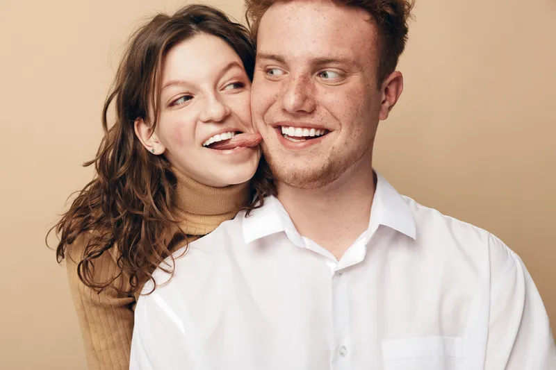Playful young couple with curly hair and red hair smiling and having fun together on beige background. Concept of love, friendship, happiness, and casual lifestyle in studio.
