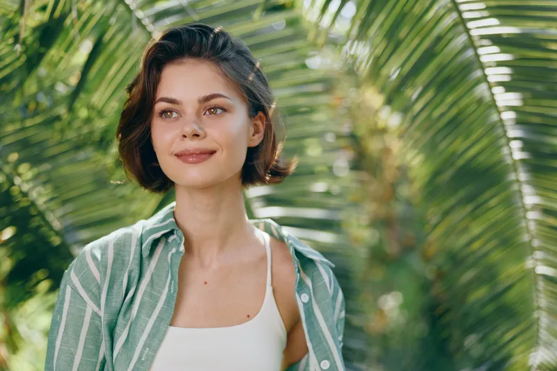 A woman portrait outdoors amid lush greenery smiles softly, wearing casual striped shirt, natural light highlighting her features, conveying calm confidence and warm approachable vibe.