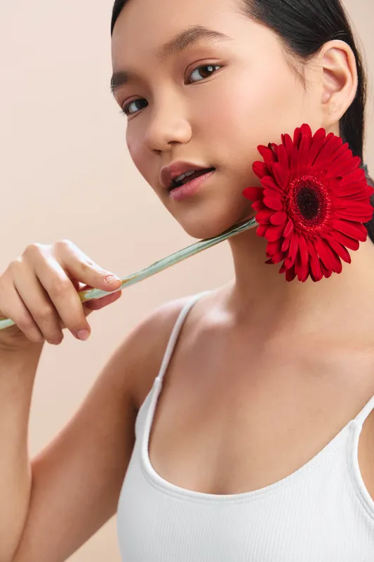 Asian woman provides a close up portrait as she holds a red flower near her face. She smiles softly, projecting warmth, grace, and serene beauty.