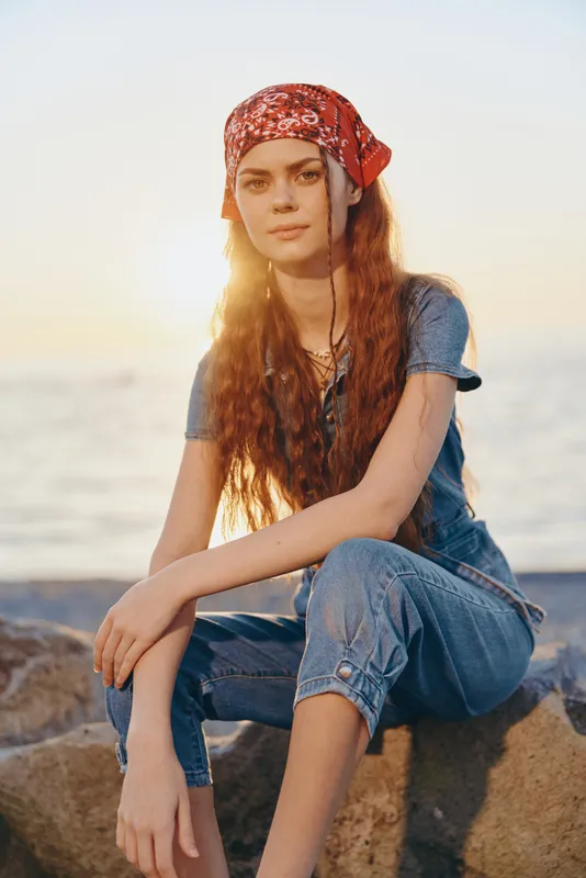 Lifestyle woman in modern boho-western denim street style with a red bandana, sitting by the seaside at sunset in warm film color tones, relaxed and natural mood.