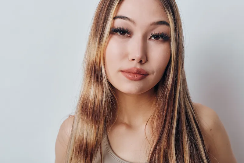 Young woman with long straight hair and natural makeup posing against light background. Female model with calm expression wearing casual top in studio portrait photography.