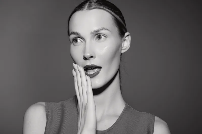 Young white woman with fair skin and slicked hair shows surprised expression touching cheek in studio portrait. Concept for emotion and beauty.