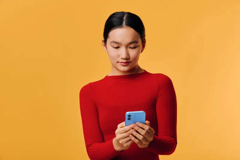 Young woman in red sweater holding and using blue smartphone for texting, browsing and social media with focused expression, isolated on orange background with casual modern style.