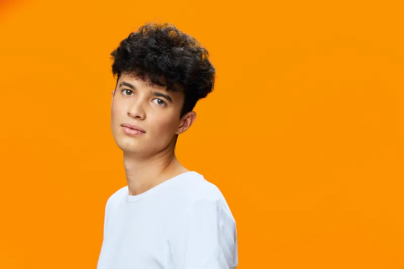 young man with curly hair wearing white t-shirt posing against bright orange background, casual style, thoughtful expression, studio portrait