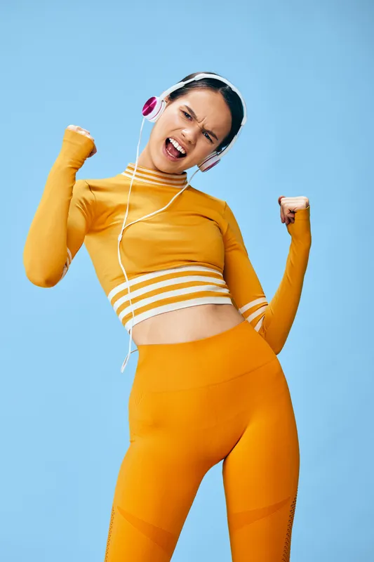Energetic woman fitness subject in bright orange activewear and crop top, wearing pink headphones, striking a celebratory workout pose with clenched fists against a clean blue studio backdrop.