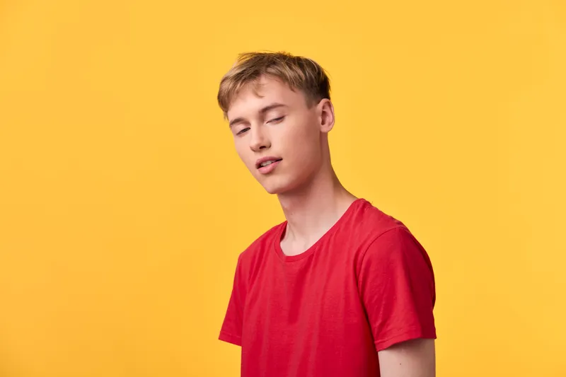 Young man in a red shirt leans with closed eyes against a vibrant yellow backdrop, conveying calm, simplicity, and relaxed style suitable for fashion and lifestyle visuals