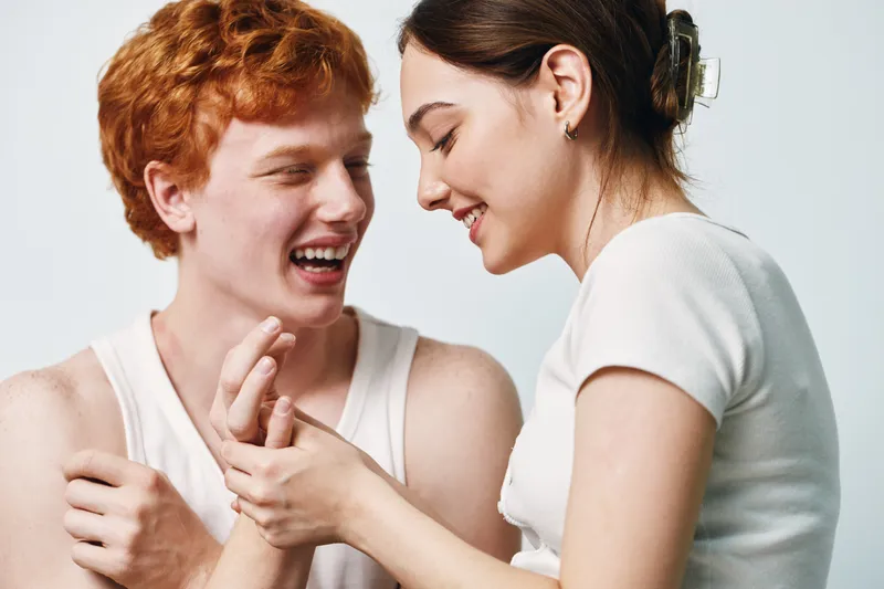 Young man and woman smiling and interacting closely in studio, casual clothing, red hair, and hair clip, friendship and joyful moment captured with white background and natural light.